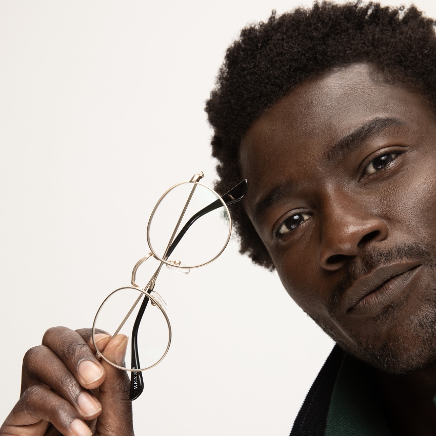 Person holding gold-rimmed round eyeglasses with black temple tips near their face.