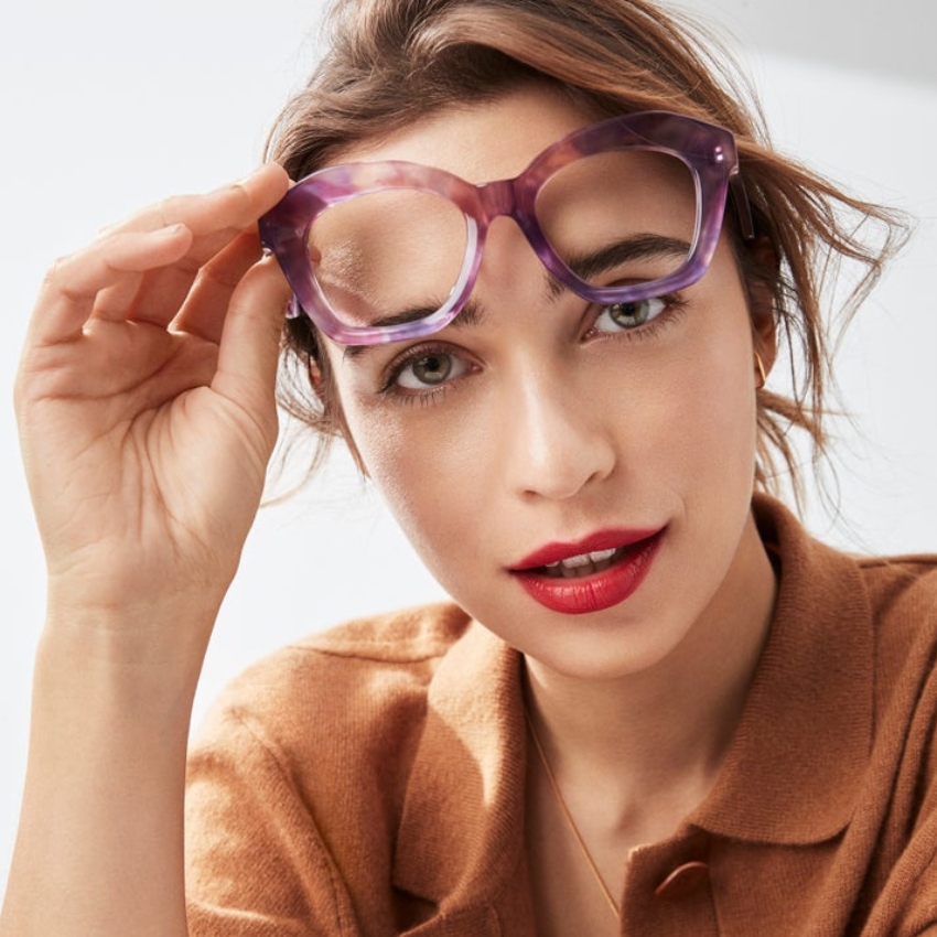 Woman wearing purple eyeglasses, slightly lifting them with her right hand. She has red lipstick on.