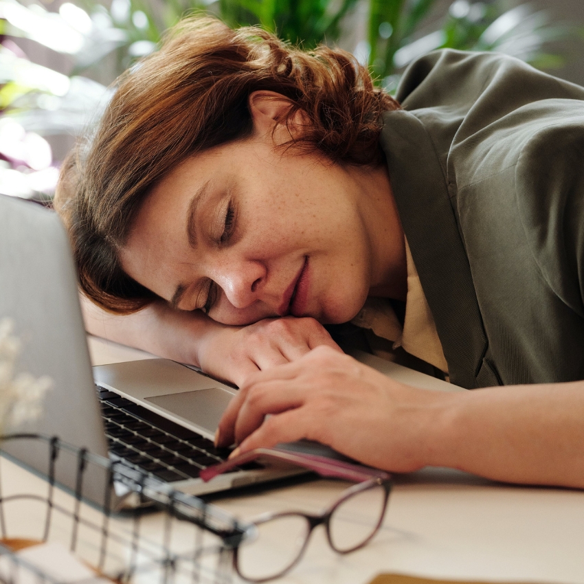 Woman resting her head on a laptop keyboard with glasses on the desk nearby.