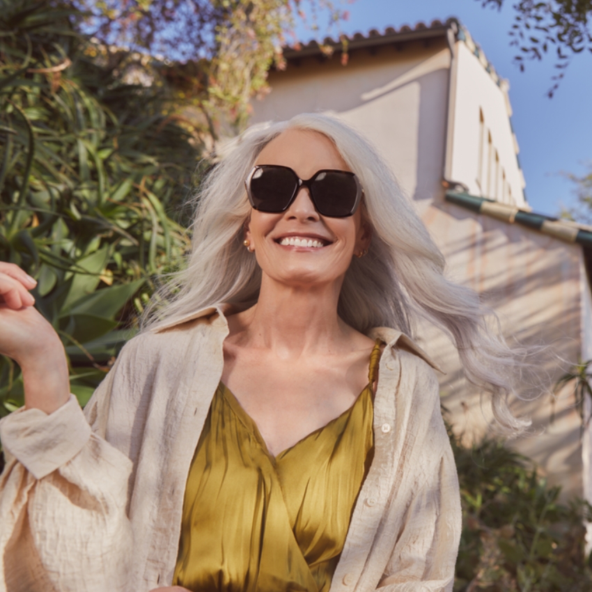 Smiling woman with long silver hair wearing oversized black sunglasses, a green dress, and a light beige shirt.