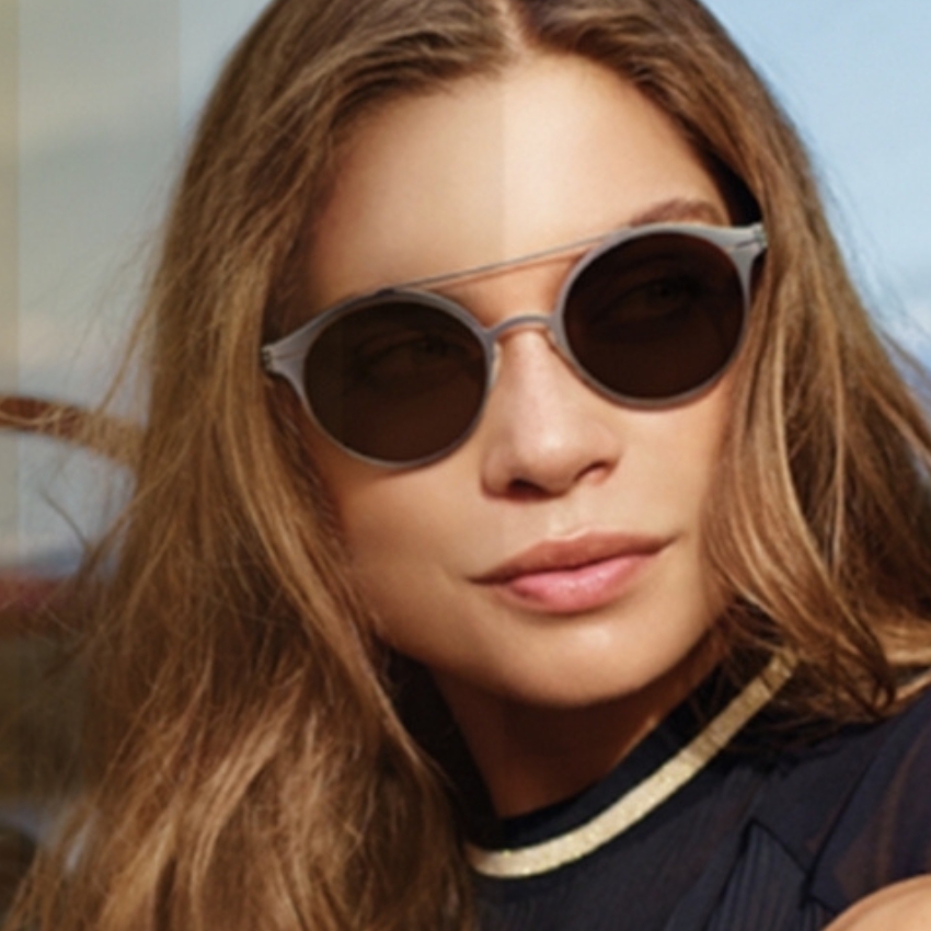 Woman wearing round, dark sunglasses with light gray frames and long, light brown hair blowing slightly.