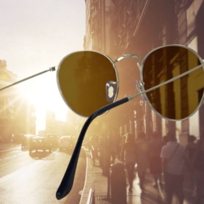 Gold-framed sunglasses with brown lenses and black temple arms, set against a sunlit city street background.