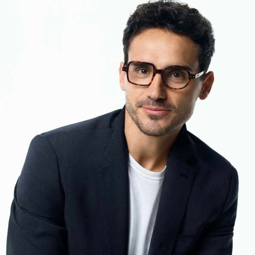 Man wearing black and brown tortoiseshell glasses, black blazer, white t-shirt.