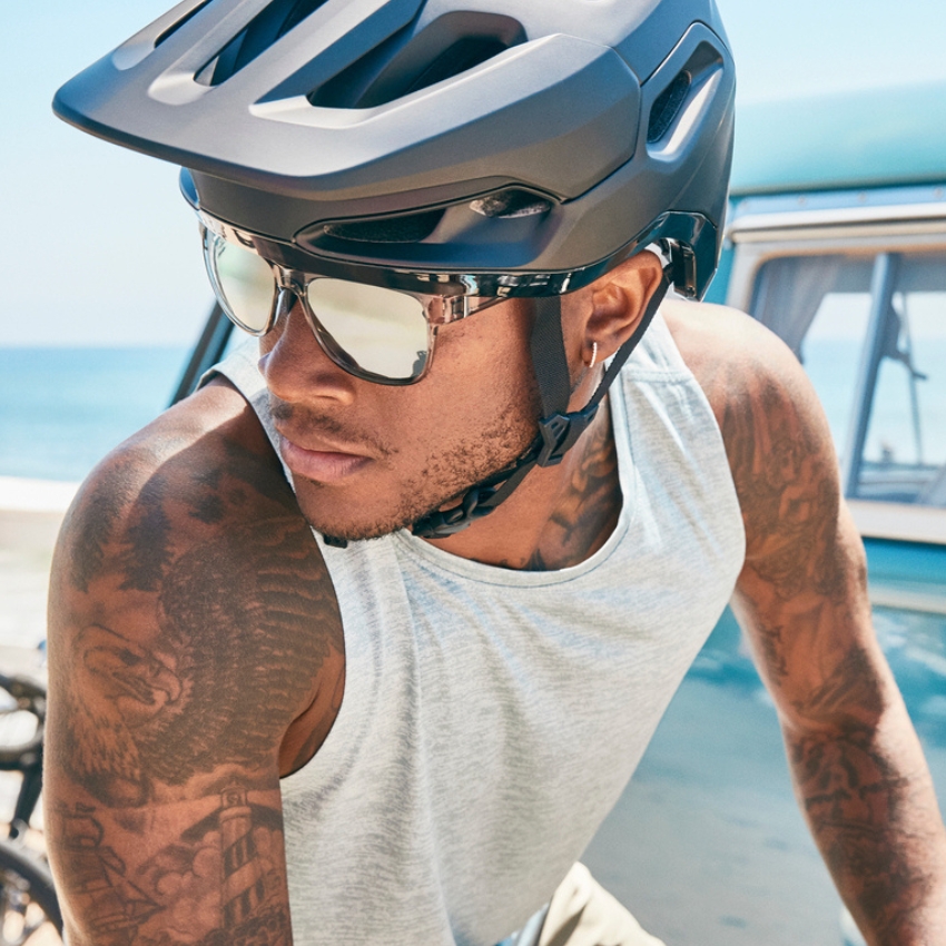 Man wearing a black bike helmet and reflective sunglasses while biking near the ocean.