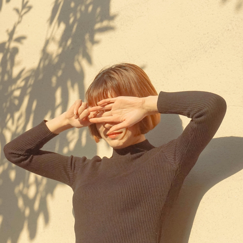 Person wearing a ribbed, black, long-sleeved shirt shielding face with hands from sunlight.