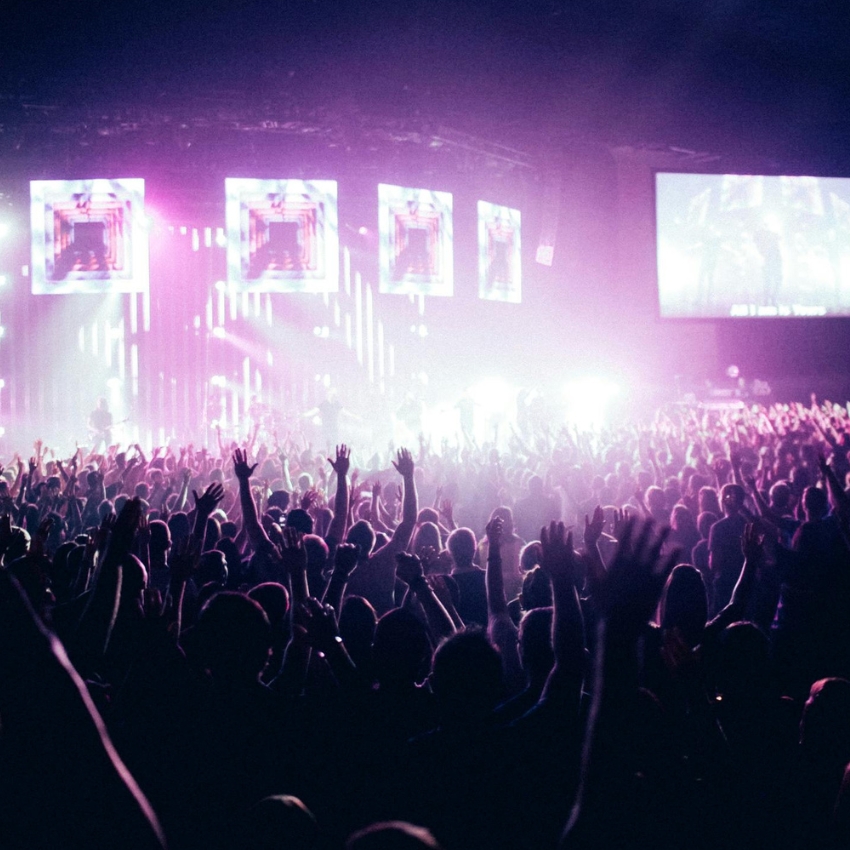 Concert audience with hands raised, stage lit with vibrant purple and white lights, screens displaying geometric designs.