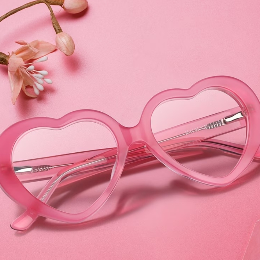 Pink heart-shaped sunglasses with clear lenses on a pink surface. Flowers are in the background.