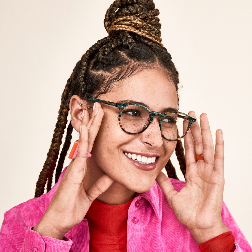 Person wearing tortoiseshell patterned eyeglasses and a pink shirt, smiling with hands near face.