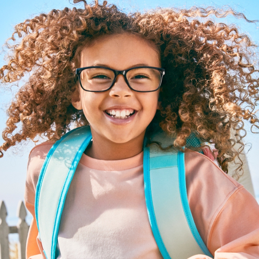 Child wearing black glasses and a light blue backpack, smiling with curly hair blowing in the wind.