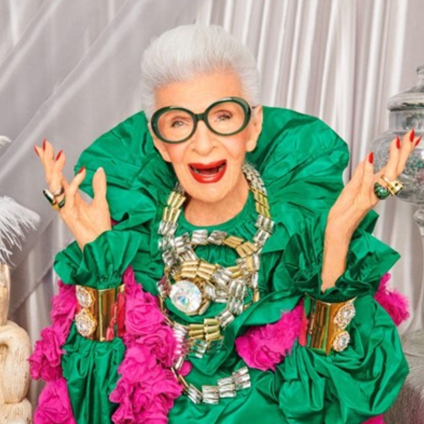 Person wearing a bright green outfit with large pink ruffles, bejeweled accessories, and green-framed glasses.