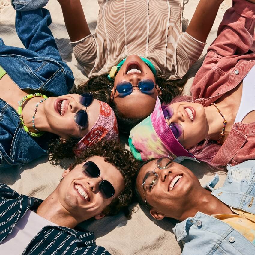 Group of five friends lying on the beach, all wearing colorful sunglasses and smiling.