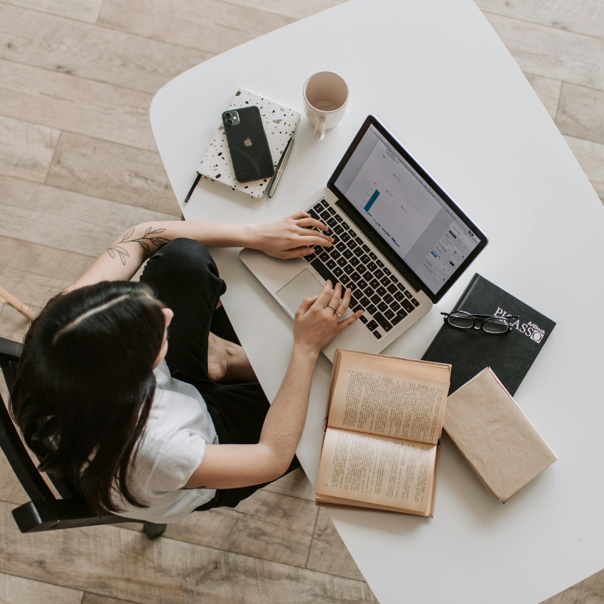 Person typing on a laptop at a white desk, with books, a notebook, a phone, glasses, and a coffee cup nearby.