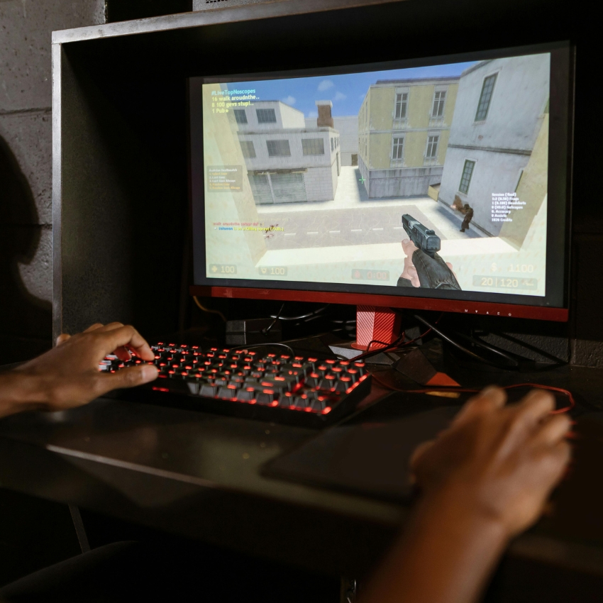 Person playing a first-person shooter game on a monitor with a mechanical keyboard illuminated with red backlight.