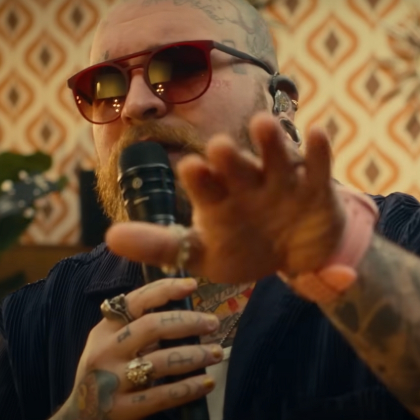 Tattooed man with red sunglasses holding a microphone and wearing multiple rings.