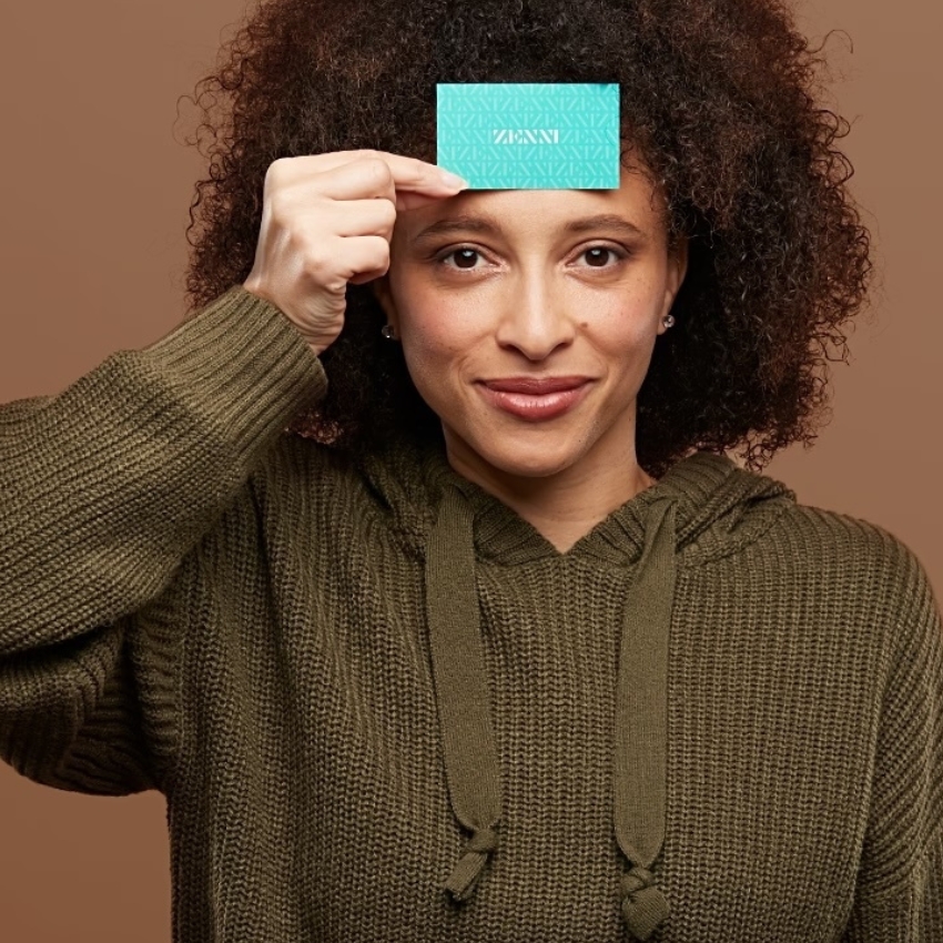 Woman holding teal Zenni card to forehead, wearing green hoodie. Text on card reads: "ZENNI."