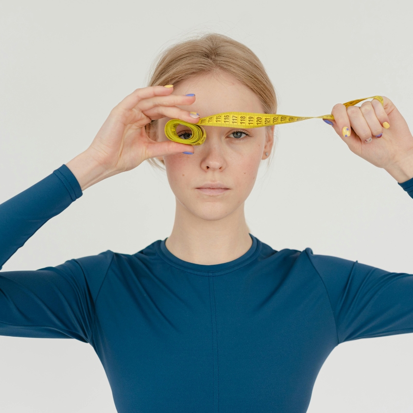 Woman in blue long-sleeve holding a rolled-up yellow tape measure, extending it across her face.