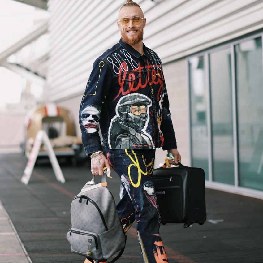 Man wearing a colorful graffiti jacket and matching trousers, carrying a gray backpack and black suitcase.