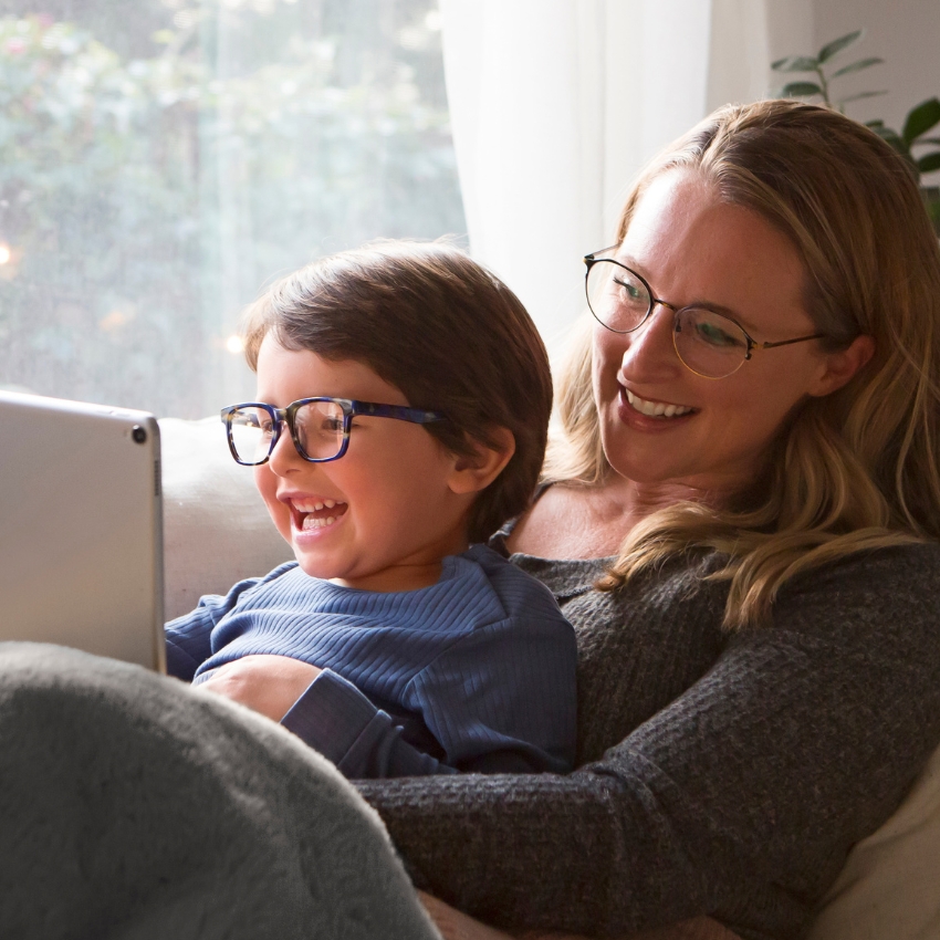 Child and woman wearing glasses, smiling and looking at a tablet screen.