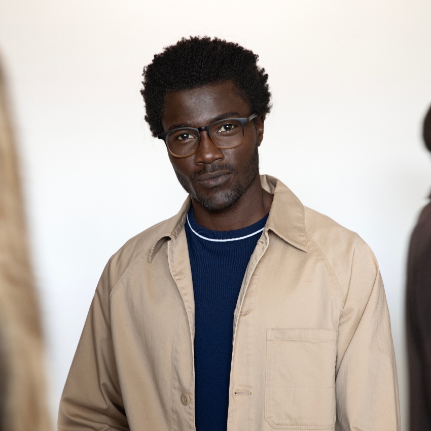 Man wearing tortoiseshell eyeglasses, a beige jacket, and a navy blue sweater.