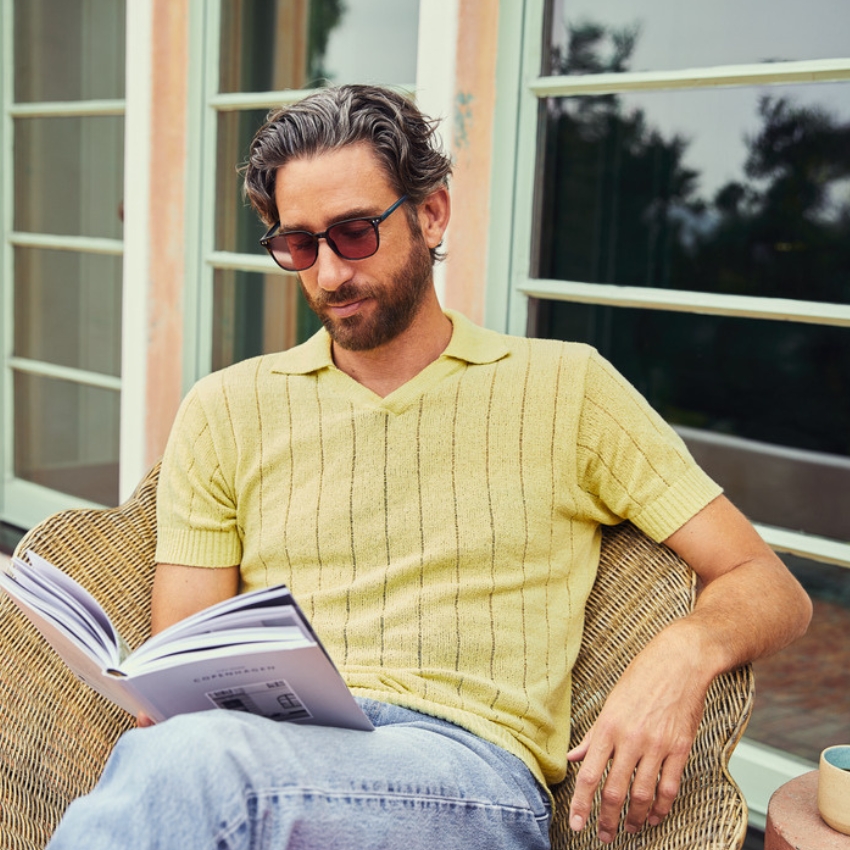 Man wearing a light yellow knit polo shirt with vertical stripes reads a book while sitting outside.