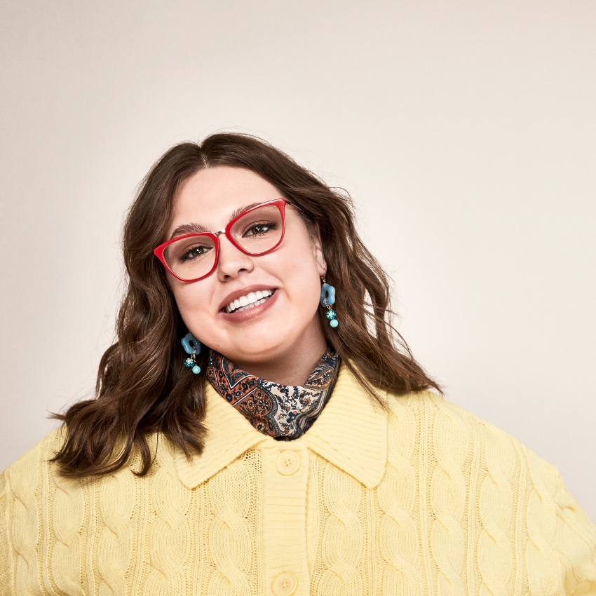 Woman in red cat-eye glasses wearing a yellow knit sweater, a patterned scarf, and blue earrings.