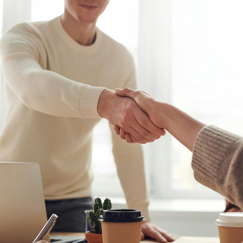 Two people shaking hands, coffee cup and small potted plant on a desk.