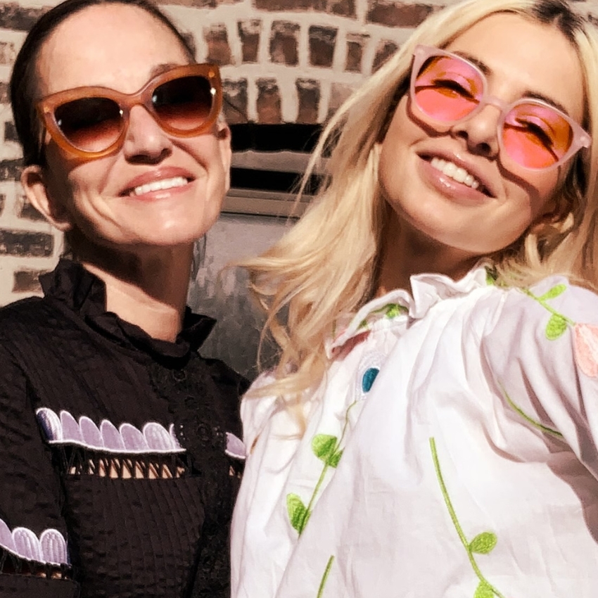 Two women wearing colorful sunglasses and detailed embroidered dresses smile brightly.