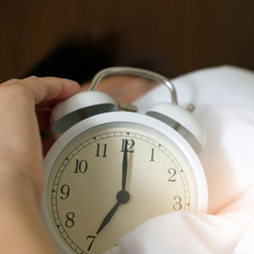 Hand holding a white analog alarm clock showing the time at 7:00.