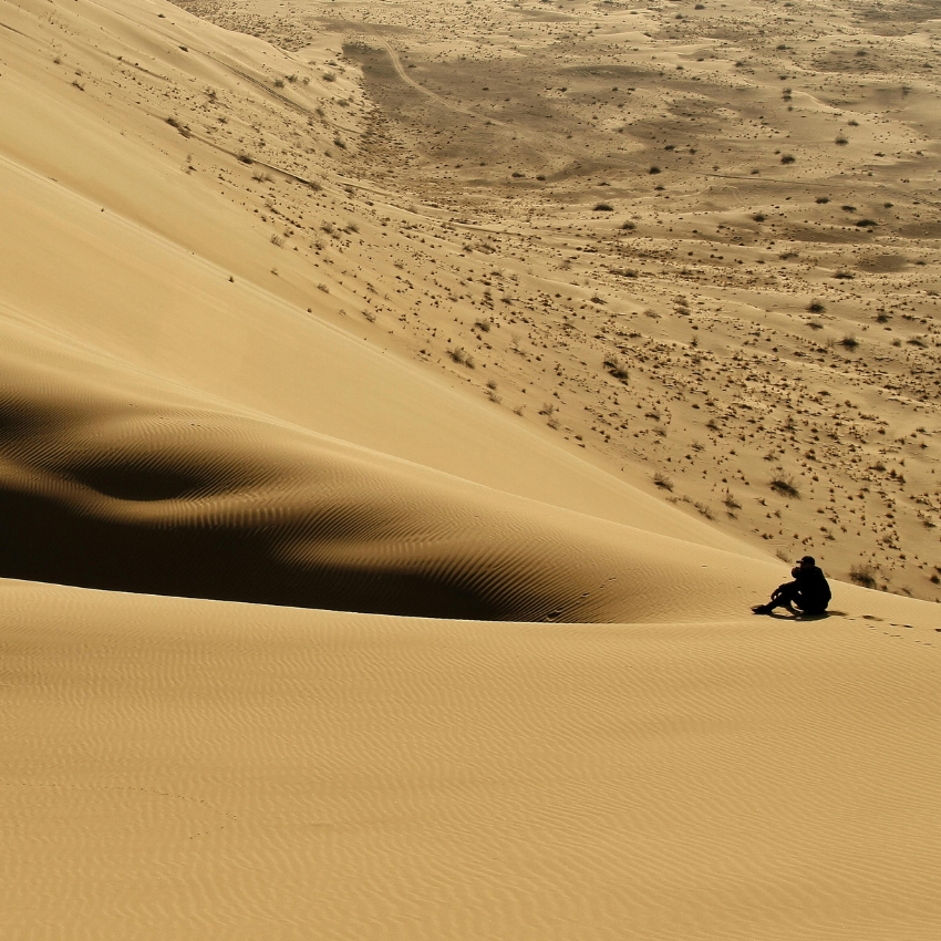 Man sitting on a large sand dune in a vast desert landscape.