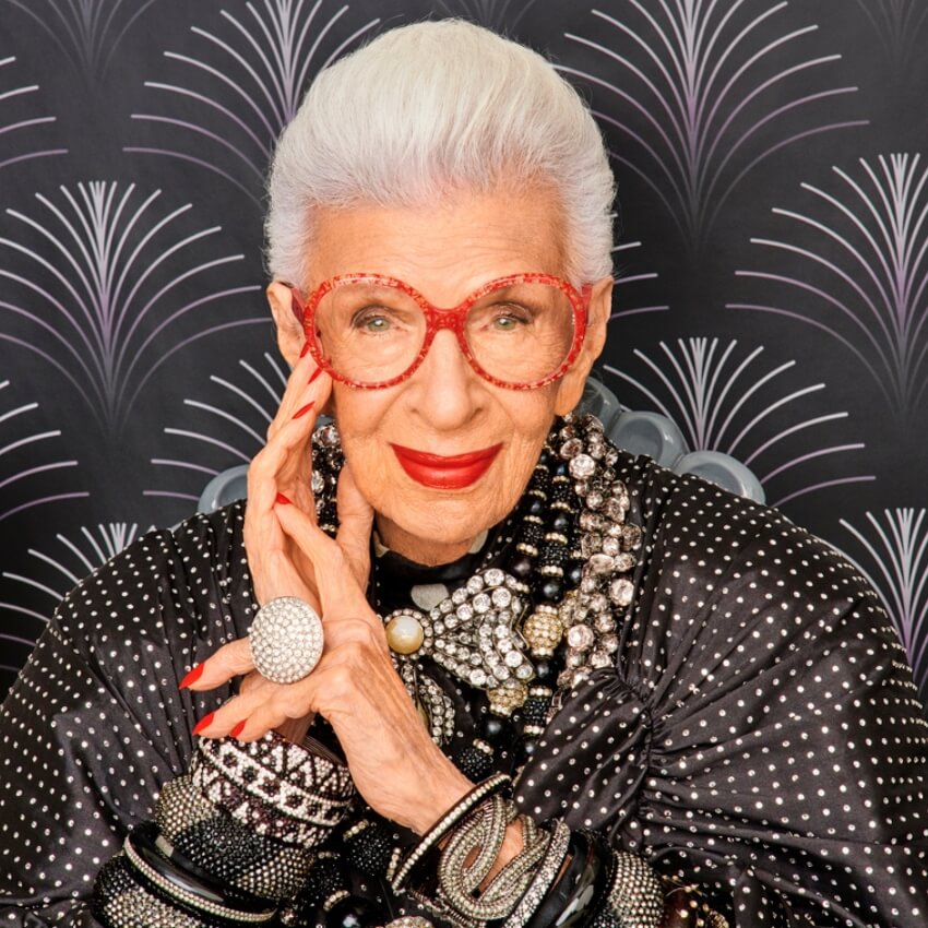 Elderly woman with white hair, red glasses, red lipstick, various beaded necklaces, and a large round ring.