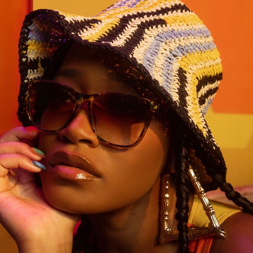 Woman wearing tortoiseshell sunglasses, a multicolored crochet bucket hat, and large bamboo-style earrings.