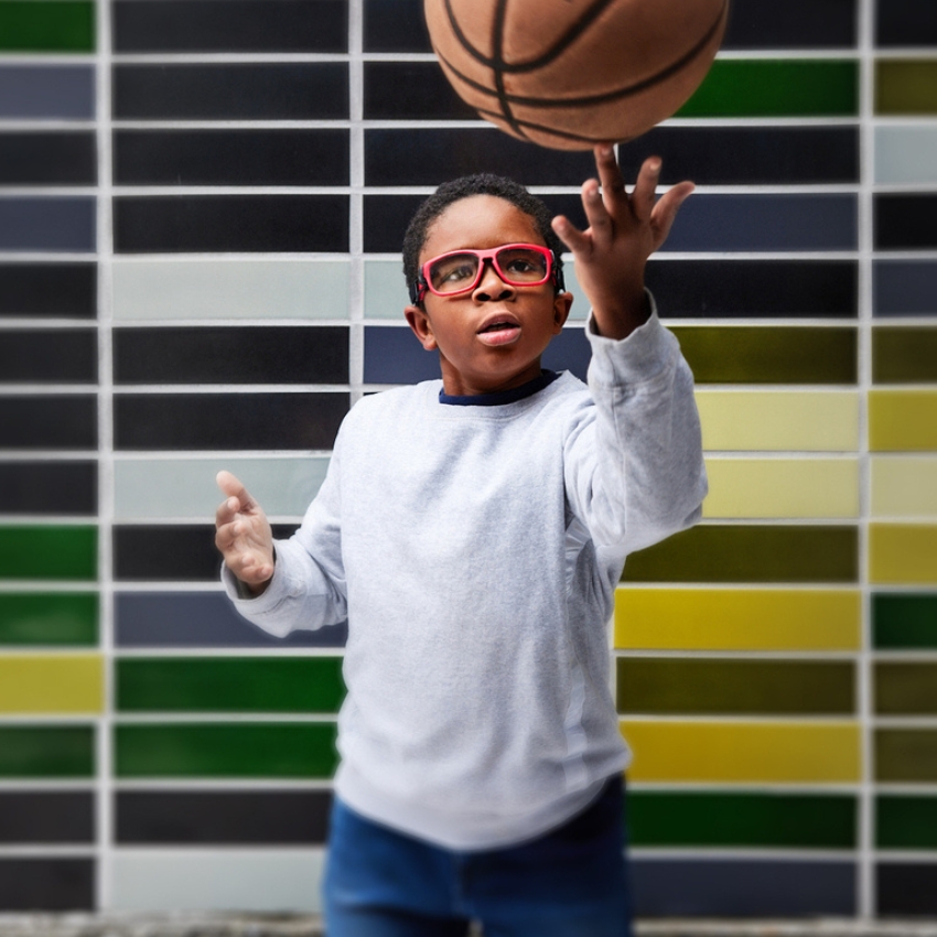 Young boy in red glasses playing with a basketball, wearing a gray sweatshirt and blue jeans.