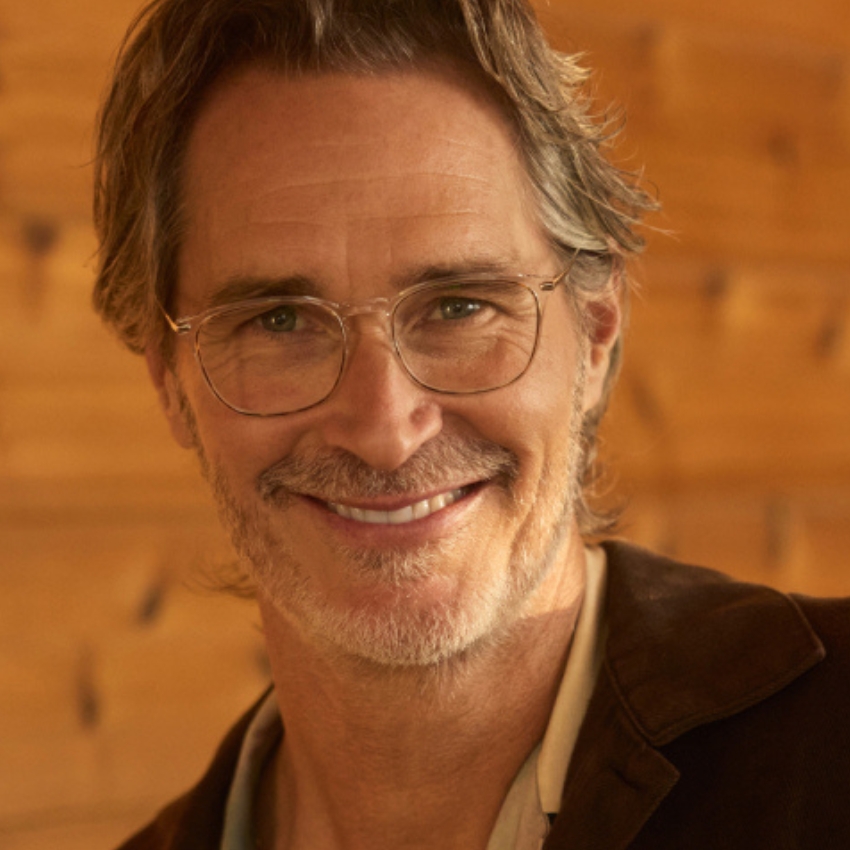 Man wearing glasses with a thoughtful smile. Brown jacket and light shirt. Warm, wooden background.