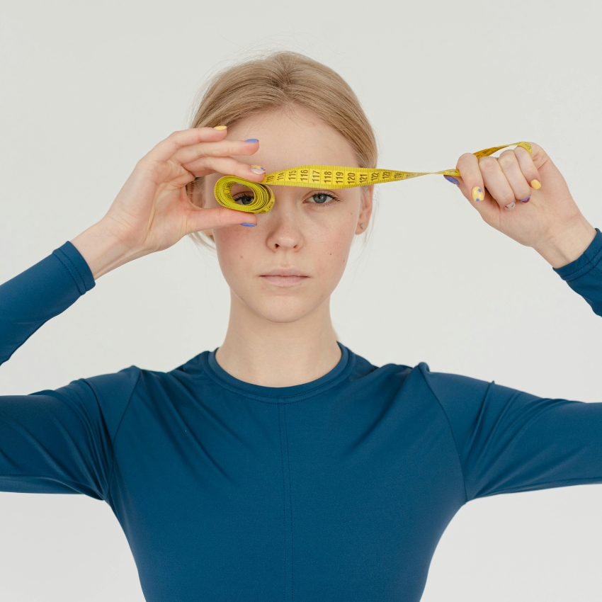 Woman in a blue long-sleeve top measuring distance with a yellow tape measure held at her eye and extended outward.