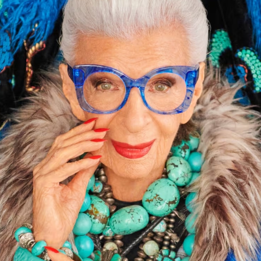 Elderly woman wearing blue glasses, chunky turquoise jewelry, and red lipstick with manicured red nails.