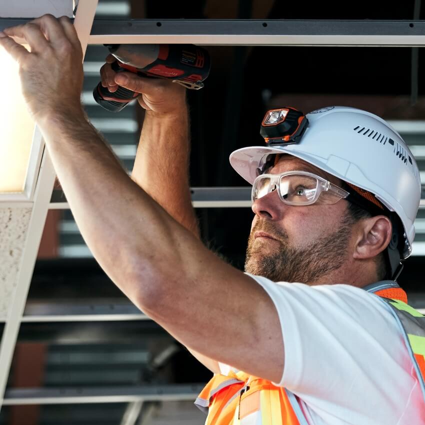 Construction worker fixing a ceiling with a drill, wearing a hard hat, safety glasses, and reflective vest.