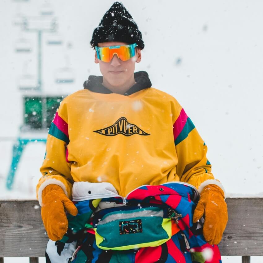 Person wearing Pit Viper yellow jacket, colorful fanny pack, orange gloves, black beanie, and reflective sunglasses.