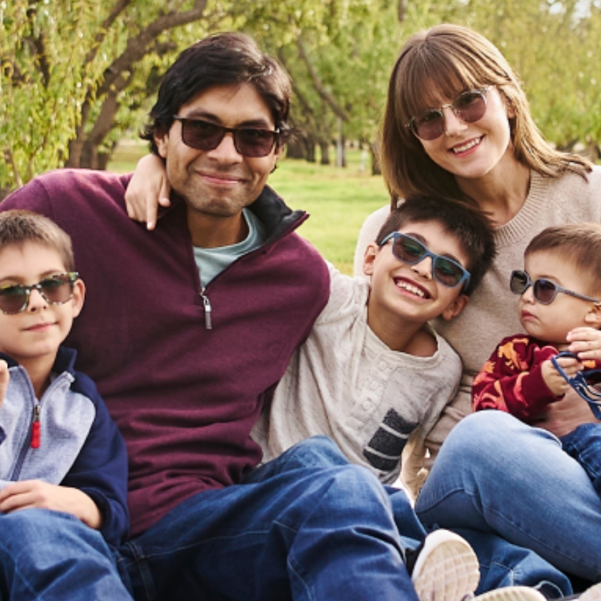 Five people wearing sunglasses and casual clothing, sitting closely together outdoors, smiling happily.