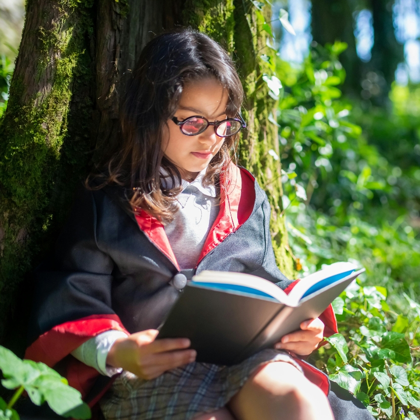 Child reading a book while wearing a black robe with red lining and glasses.