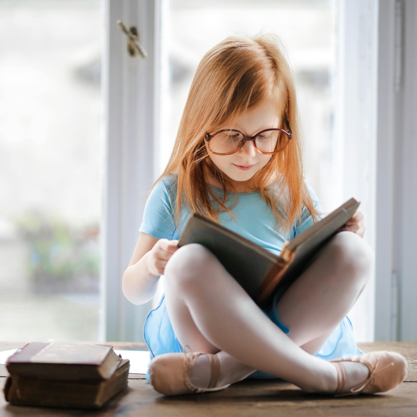 Young girl with glasses reading a book, sitting cross-legged in a light blue dress and ballet shoes.