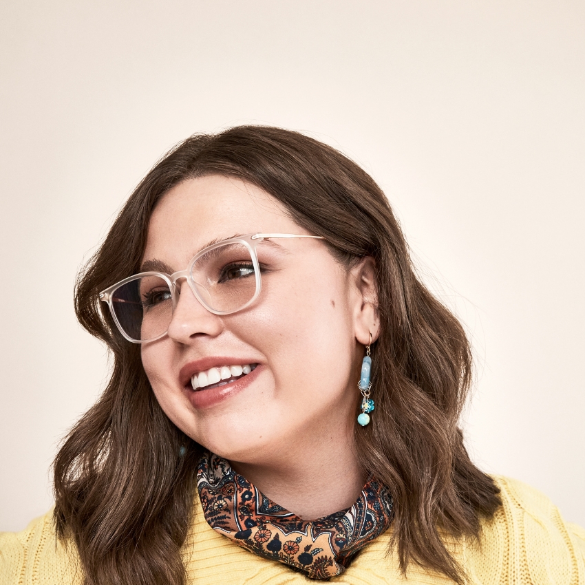 Woman wearing clear-frame glasses, blue and turquoise drop earrings, and a paisley patterned neck scarf.