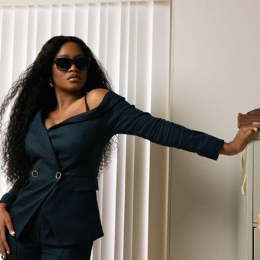 Woman in navy blazer and pants, one shoulder exposed, wearing black sunglasses and leaning on a shelf.