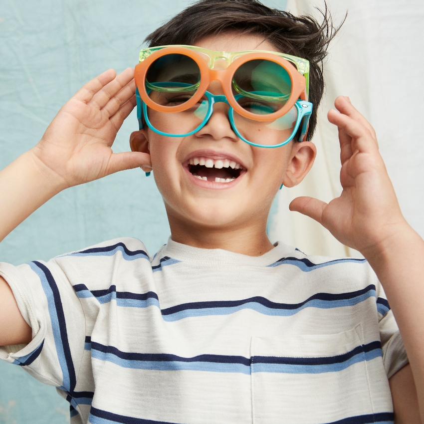 Child wearing oversized orange and blue sunglasses, smiling joyfully while holding the glasses.