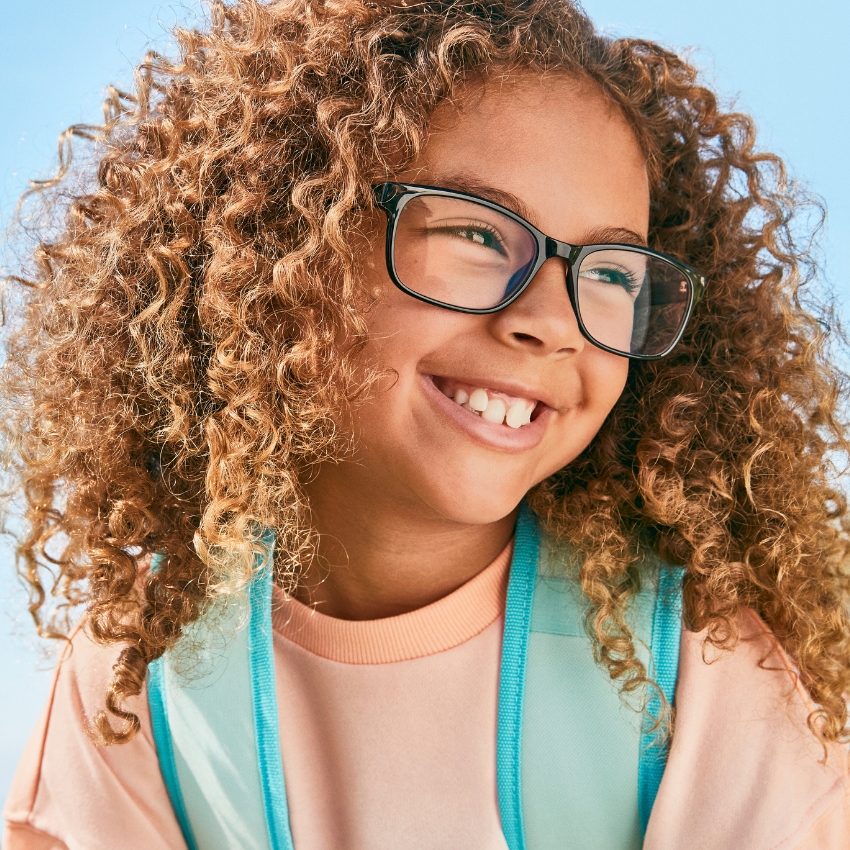 Smiling child with curly hair wearing black-rimmed glasses and a pastel pink shirt with a teal backpack.