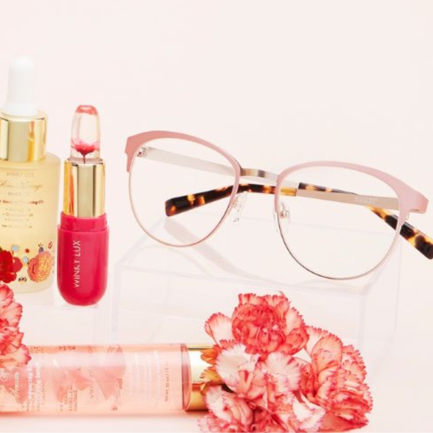 Winky Lux glasses, serum bottle, lipstick, and spray surrounded by pink flowers.