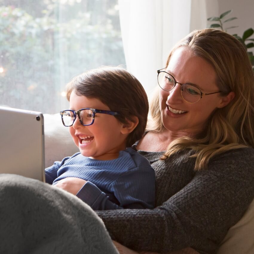 A child and an adult wearing glasses, smiling and looking at a tablet screen.