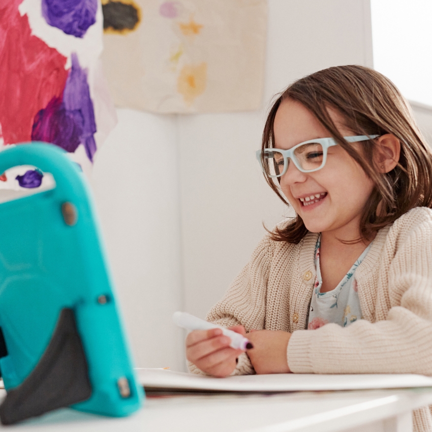 Child wearing glasses using a teal tablet while holding a marker and smiling.