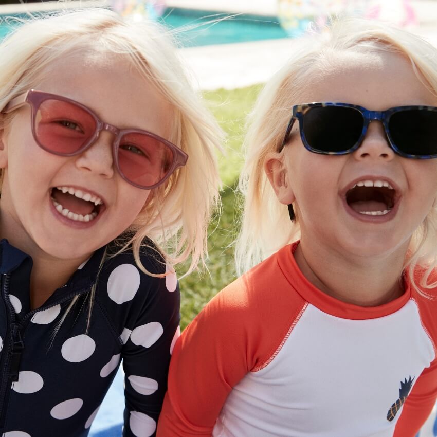 Children wearing polarized sunglasses, left one with red lenses, right one with dark blue lenses, smiling.