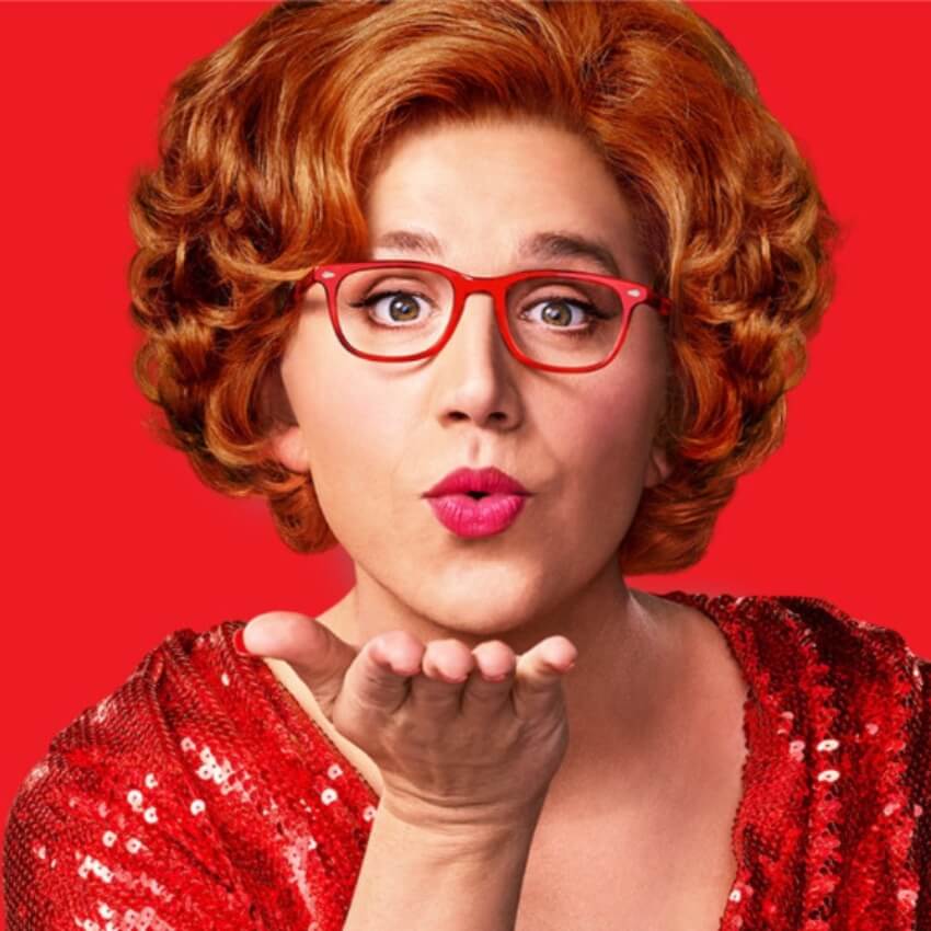 Person with red glasses and red lipstick blowing a kiss, wearing a red sequined outfit against a red background.