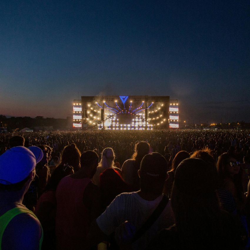 Large crowd at an outdoor concert at dusk, with a brightly lit stage in the background.